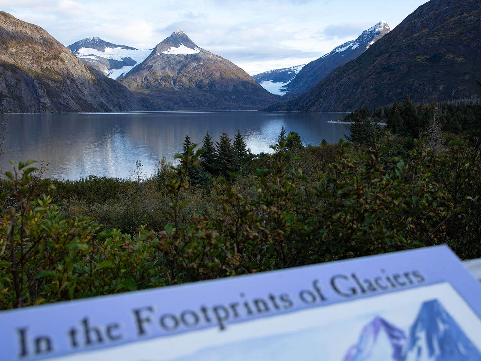 A portion of the "In the Footprints of Glaciers" book cover in frame in front of a view of the lake in the Portage Valley